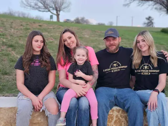 Family of five smiling, sitting together outside on a concrete ledge.
