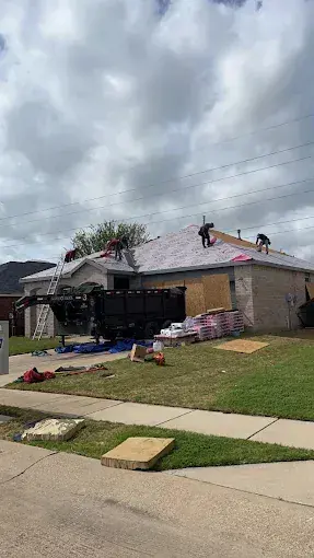 Roofers working on a house roof. Dumpster and supplies in the yard, cloudy sky overhead.