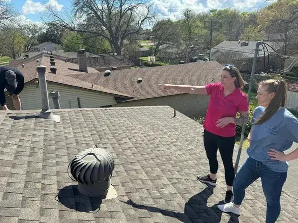 Three people on a brown shingle roof, one pointing, on a sunny day