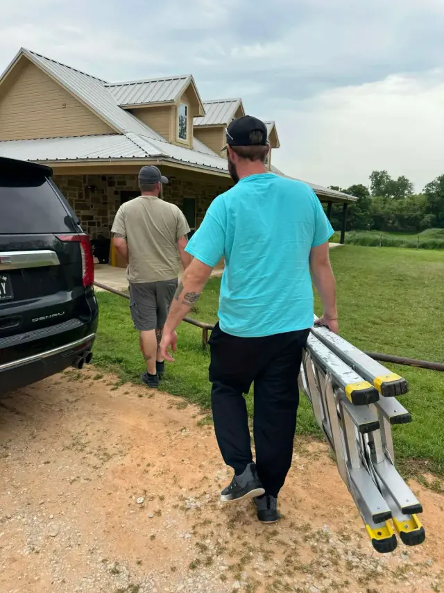 Two men walk towards a house, one carrying a folded ladder. A black SUV sits in the driveway.