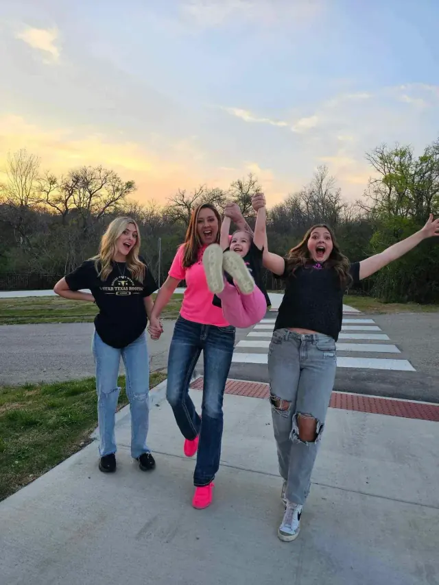 Four people smiling and playing on a paved area, under a sunset sky. One person is holding a child up by their hands.