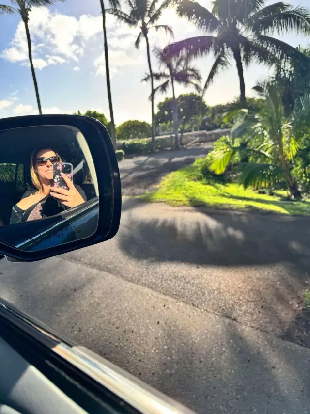 Woman in sunglasses taking a selfie reflected in car's side mirror, driving on a sunny road lined with palm trees.