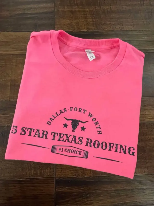 Pink t-shirt with black text: "5 Star Texas Roofing" and Dallas-Fort Worth, plus a longhorn. Folded on wood.