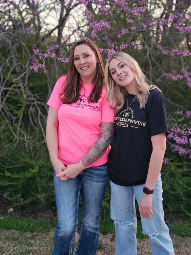 Two women smiling, posing outdoors by a bush with pink flowers. One in a pink shirt, the other in black.