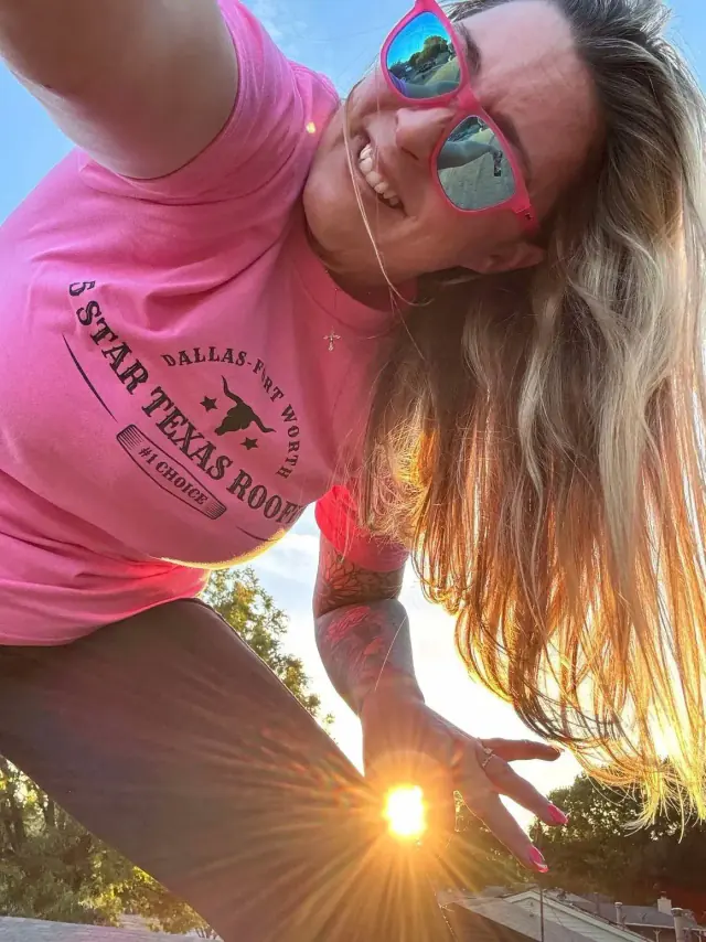 Woman in pink shirt and sunglasses holds the sun, smiling at the camera.
