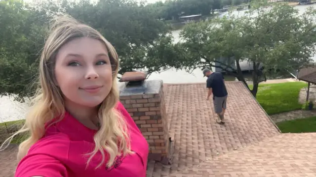 Woman in pink shirt takes a selfie on a brick roof; man walks nearby. Trees and water in background.