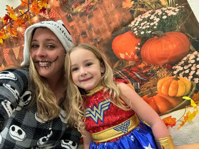 Woman and child in Halloween costumes smile in front of a pumpkin backdrop.