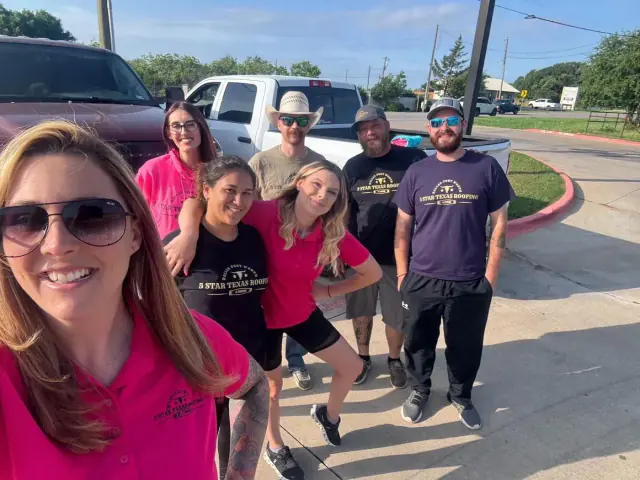 Group of people smiling, posing near cars on a sunny day; some wearing t-shirts with a logo.