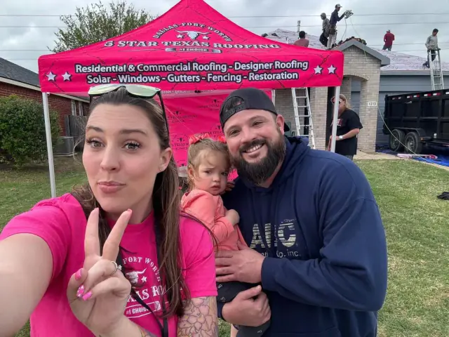 Family selfie in front of a roofing business tent; woman making peace sign, man holding child, roofers working on a roof.