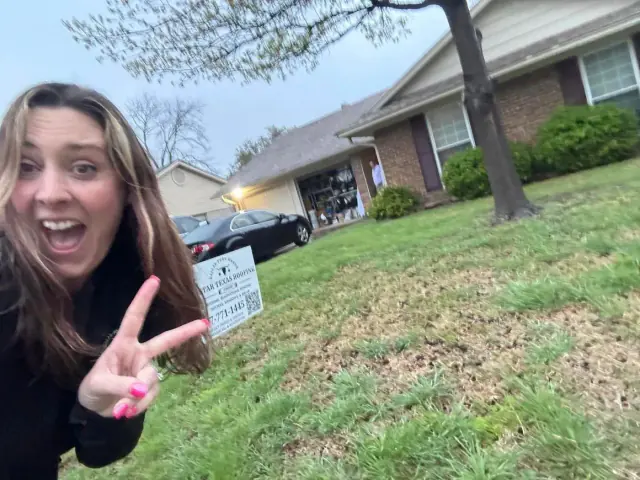 Woman making peace sign outside a house with a car in the driveway and sign.