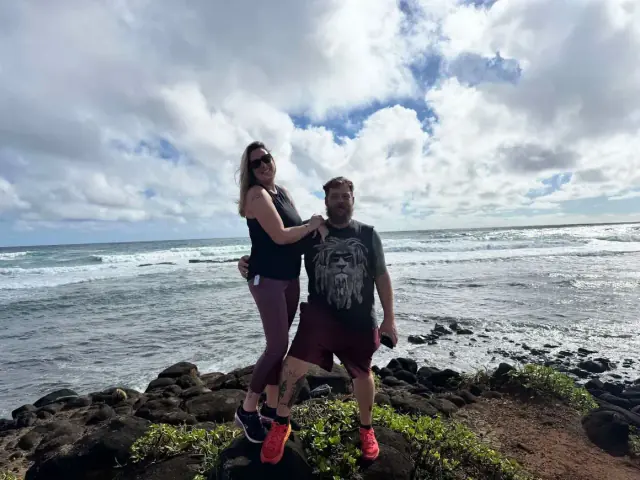 Woman stands above man by the ocean; she rests her hand on his shoulder. Cloudy sky.