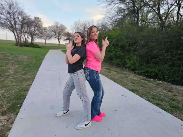 Two people stand back to back on a paved path, both pointing fingers up, in a park.