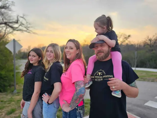 Family posing outdoors at sunset; daughter on father’s shoulders, two others standing next to them.