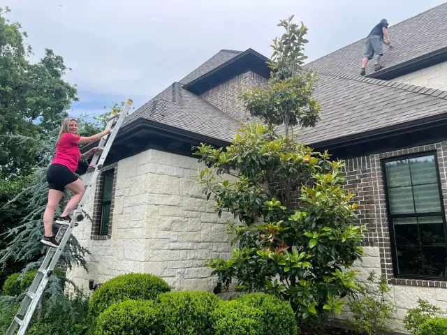 Woman on ladder reaching toward roof while a person works on the roof of a house.