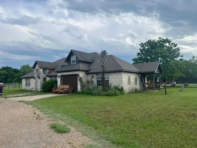 A weathered, two-story house with a lawn under a cloudy sky. A driveway leads to the house.