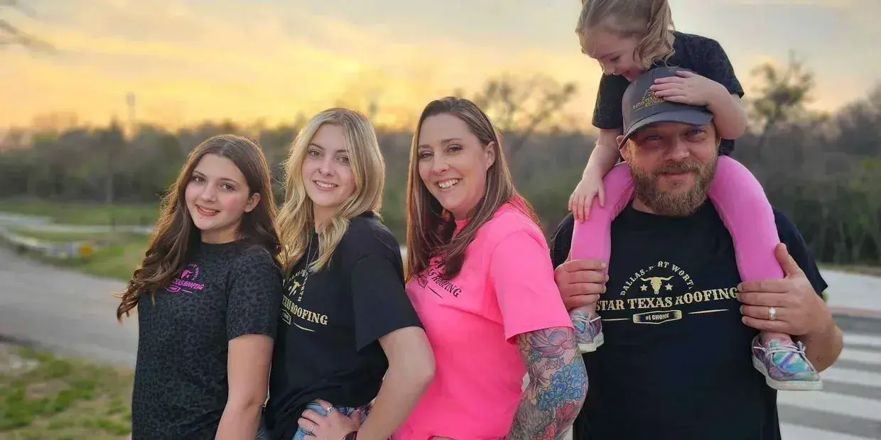 Family of five smiling, two daughters, two women, man holding child on shoulders, sunset background.