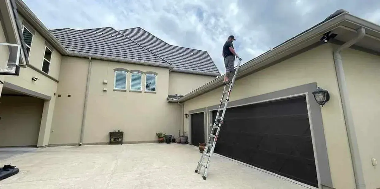 Person on a ladder cleaning gutters on a beige house with a dark garage door, cloudy sky.