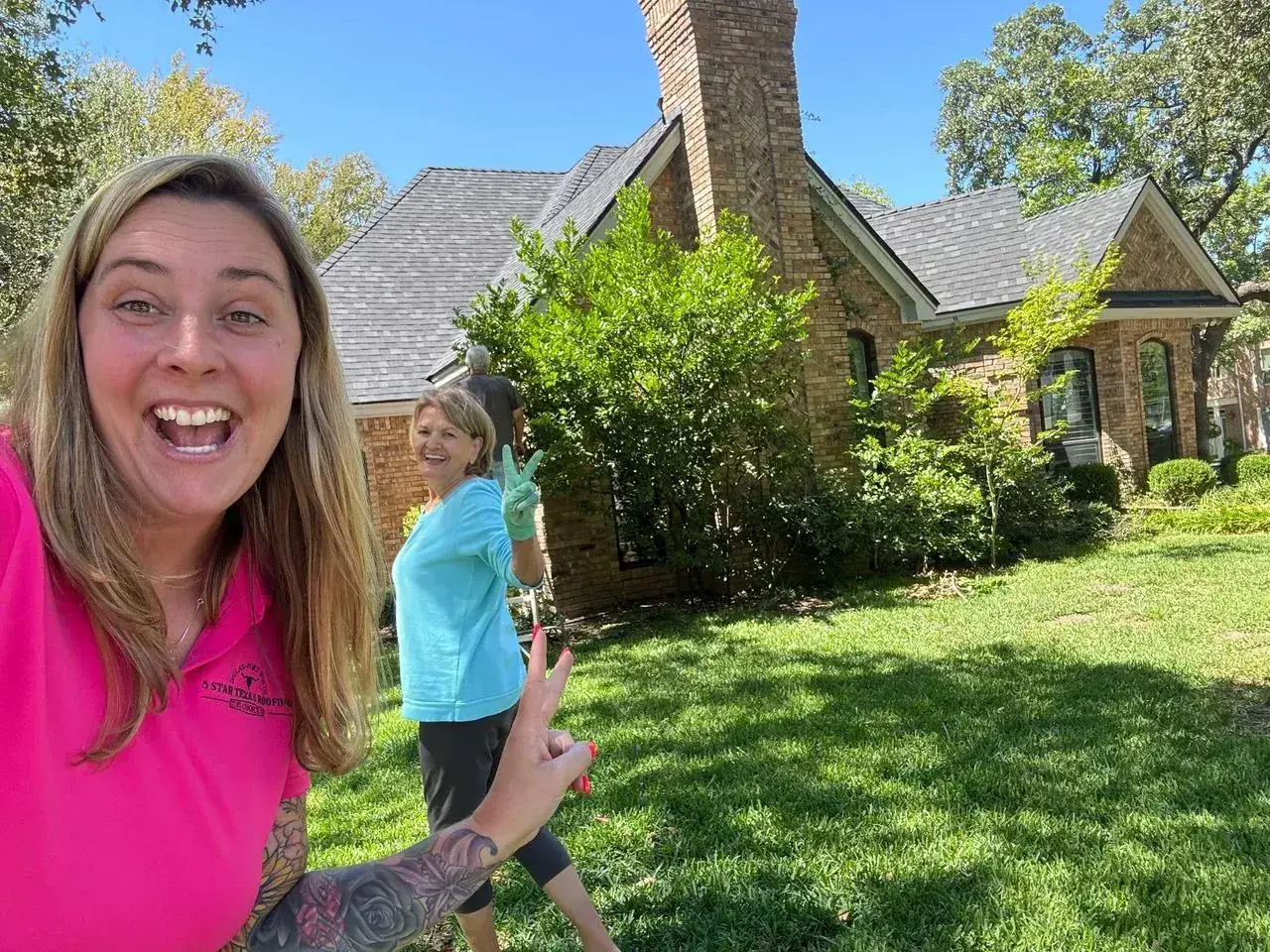 Woman in pink shirt smiles, pointing at a house with a woman in a blue shirt standing in front. Sunny day.