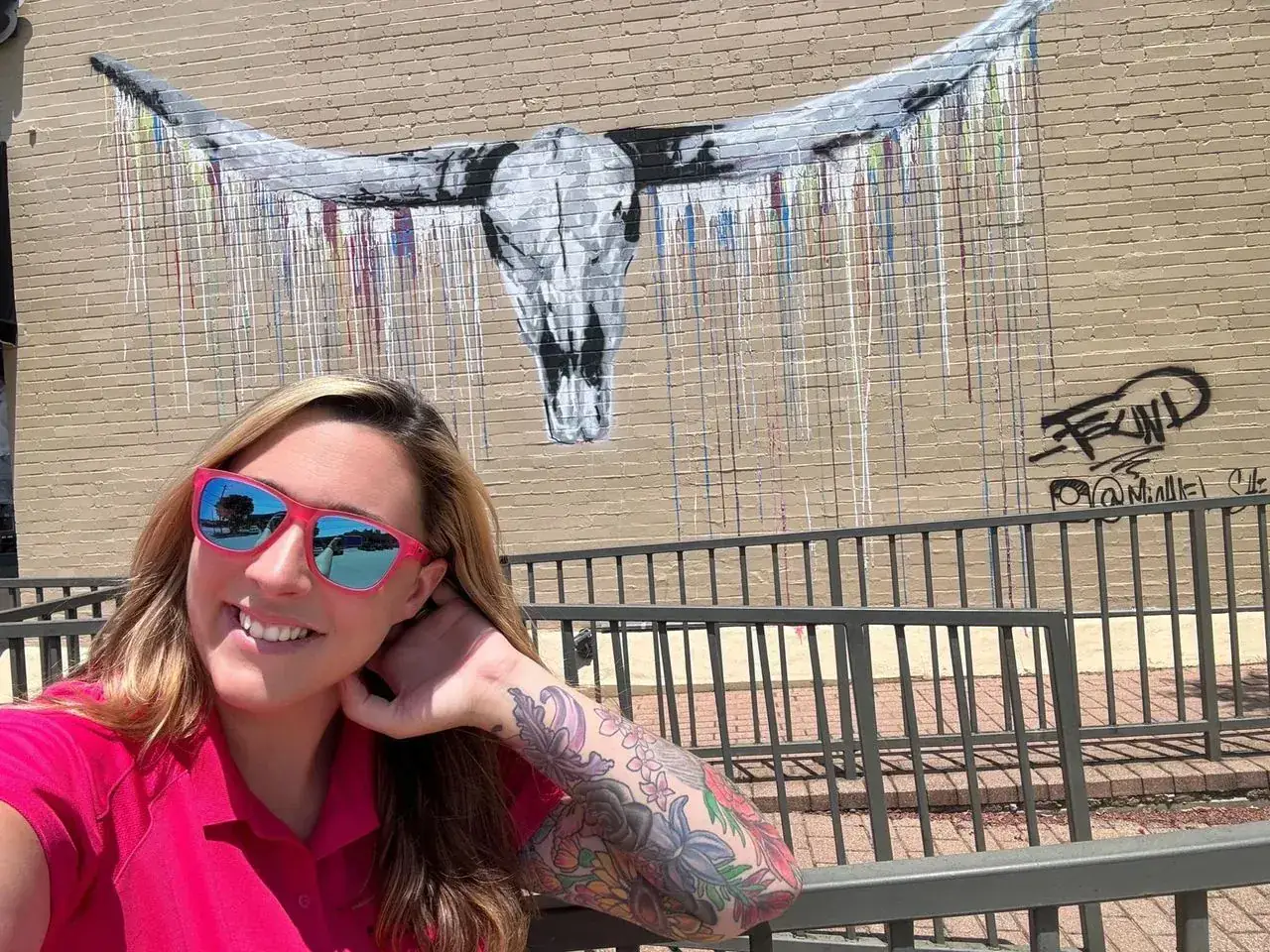 Woman in pink shirt and sunglasses smiles at the camera, posing in front of a longhorn skull mural.