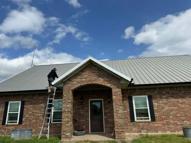 Person on a ladder on a roof, inspecting or working. Red brick house, blue sky with clouds.