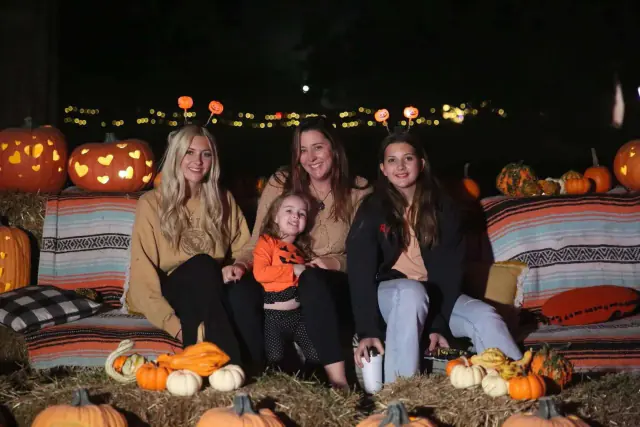 Four people sit on hay bales surrounded by pumpkins at night.