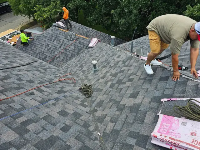 Roofers installing shingles on a multi-sloped roof. One man working close-up, others further away. Cloudy sky.
