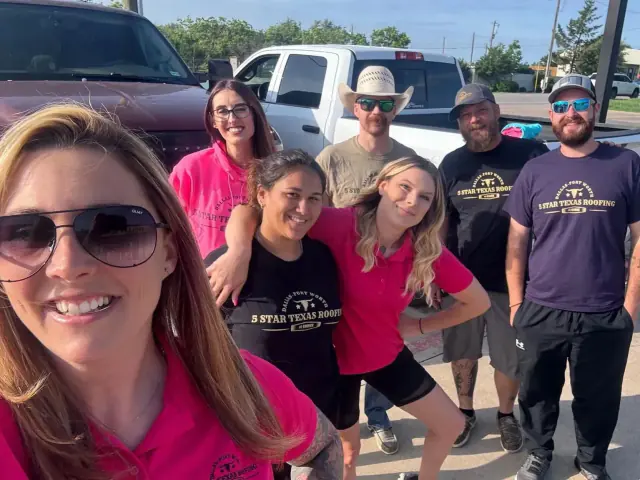 Group of people smiling near vehicles on a sunny day. Some wear matching shirts.