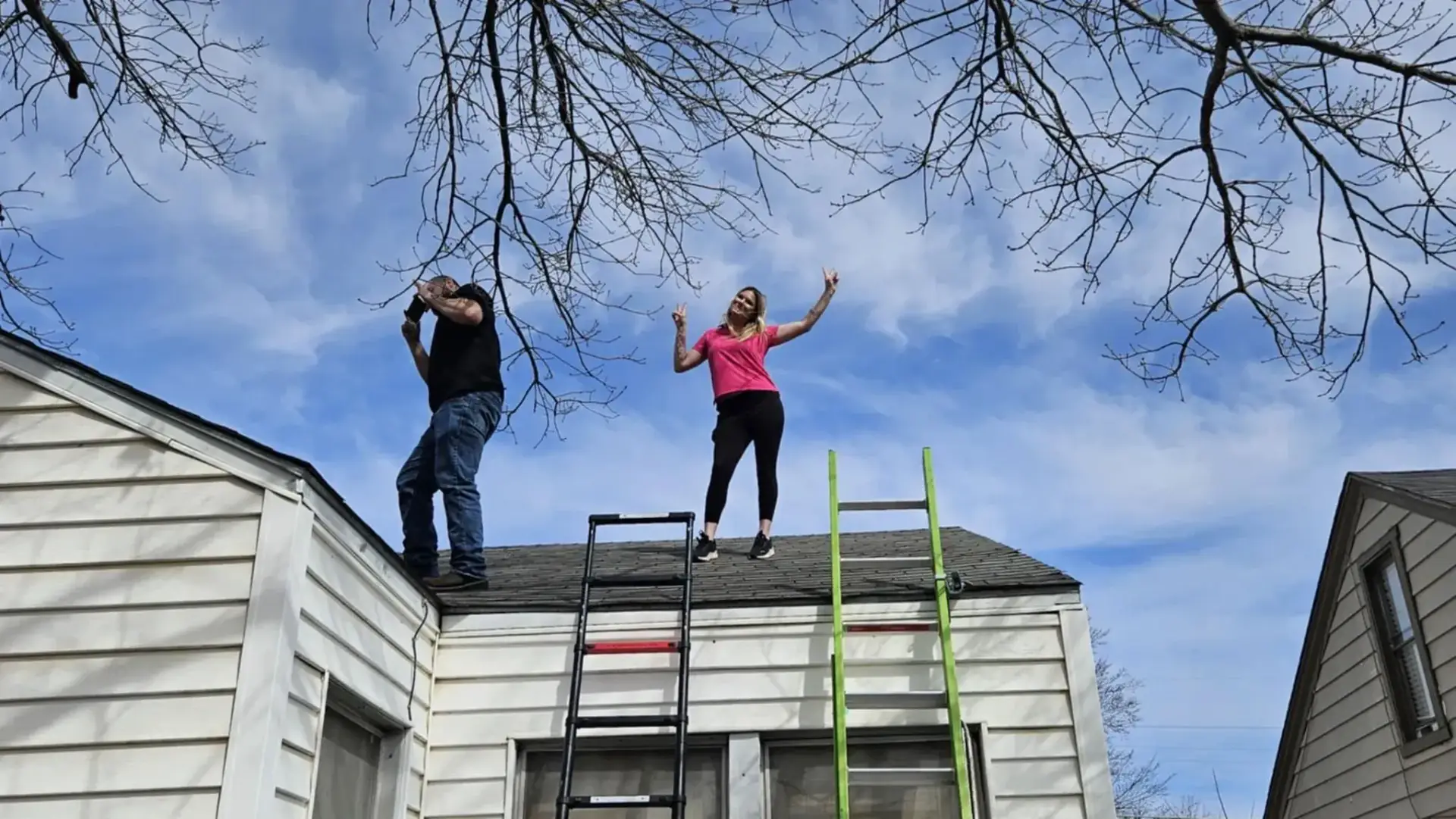 Two people standing on a rooftop under a blue sky, one raises arms, and the other points a camera.