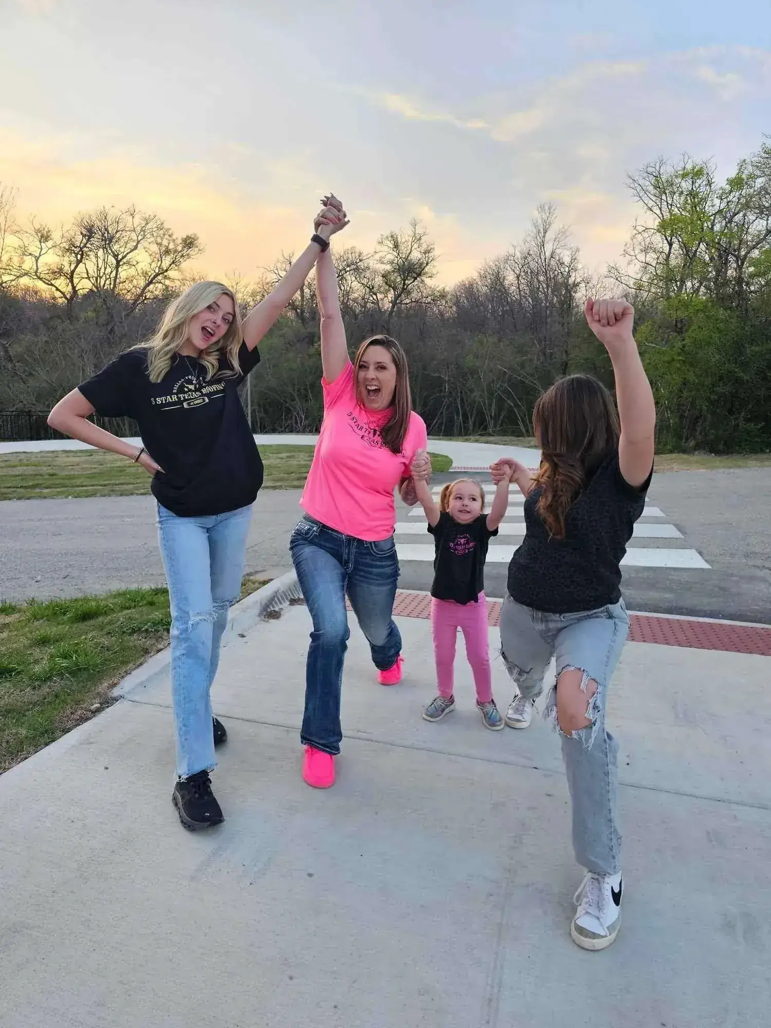 Four people celebrating outdoors, arms raised. One in pink shirt, others in black shirts and jeans.