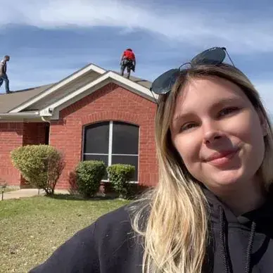 Woman smiles in front of a brick house; roofers are on the roof. Sunglasses are on her head.
