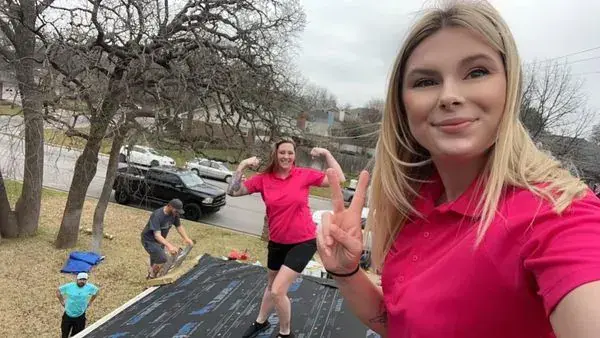 Two women on a rooftop take a selfie, another flexes, two men are working. Trees, a street, cars.