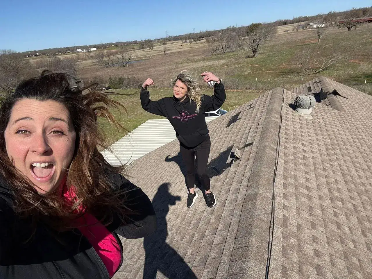 Two women on a shingle roof in open countryside, one flexing for the camera
