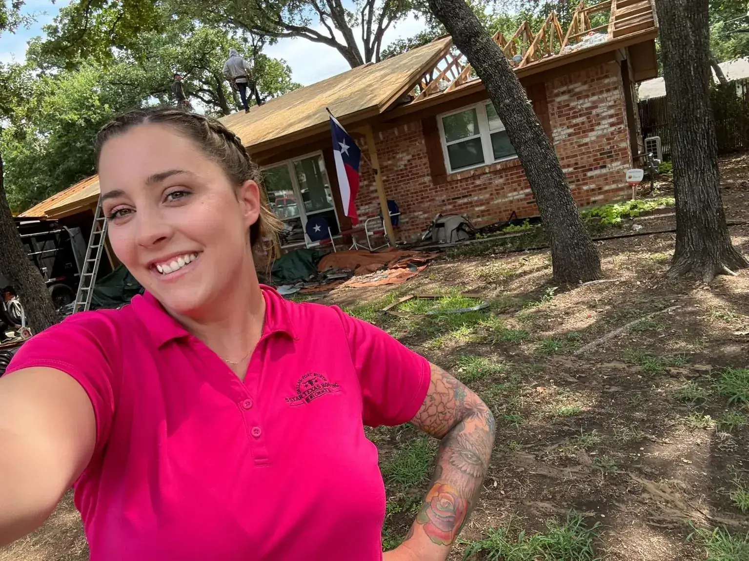 Woman in a pink shirt taking a selfie in front of a house with a roof tear-off underway
